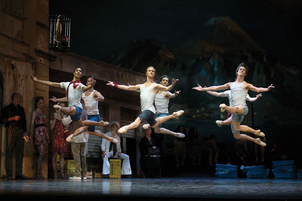 Six male dancers dressed in denim shorts and white tank tops leap in a grand jet\u00e9 en avant onstage