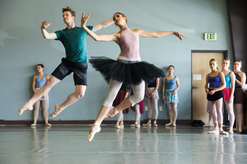 Two dancers in rehearsal clothes jump in jet\u00e9s in class with other dancers standing behind them