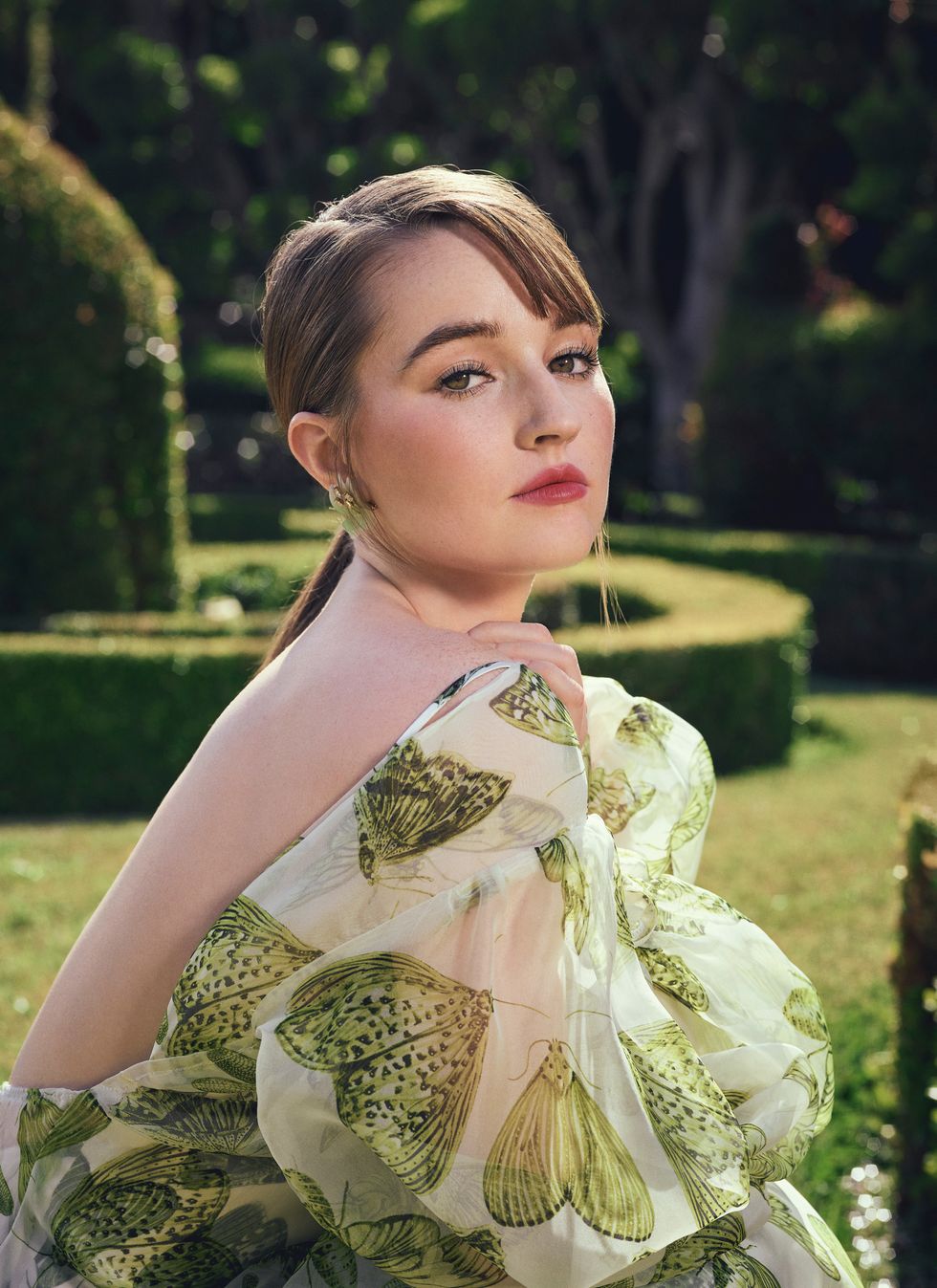 Actress Kaitlyn Dever wears a flowing butterfly print dress in a groomed topiary garden.