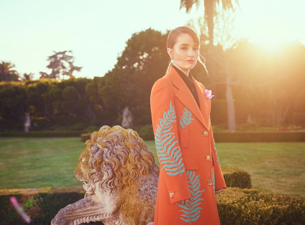 Actress Kaitlyn Dever stands next to a lion statue wearing an orange overcoat.