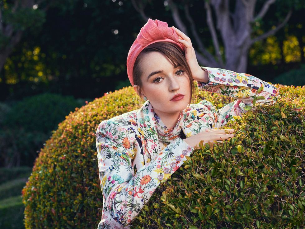 Actress Kaitlyn Dever Wears a headband with a flower print top while leaning against a topiary bush.