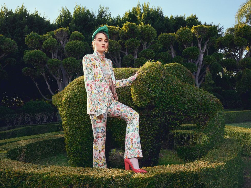 Actress Kaitlyn Dever Wears a headband with a flower print top while leaning against a topiary elephant.