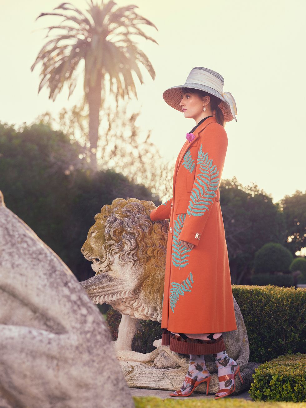 Actress Kaitlyn Dever stands next to a lion statue wearing an orange overcoat.