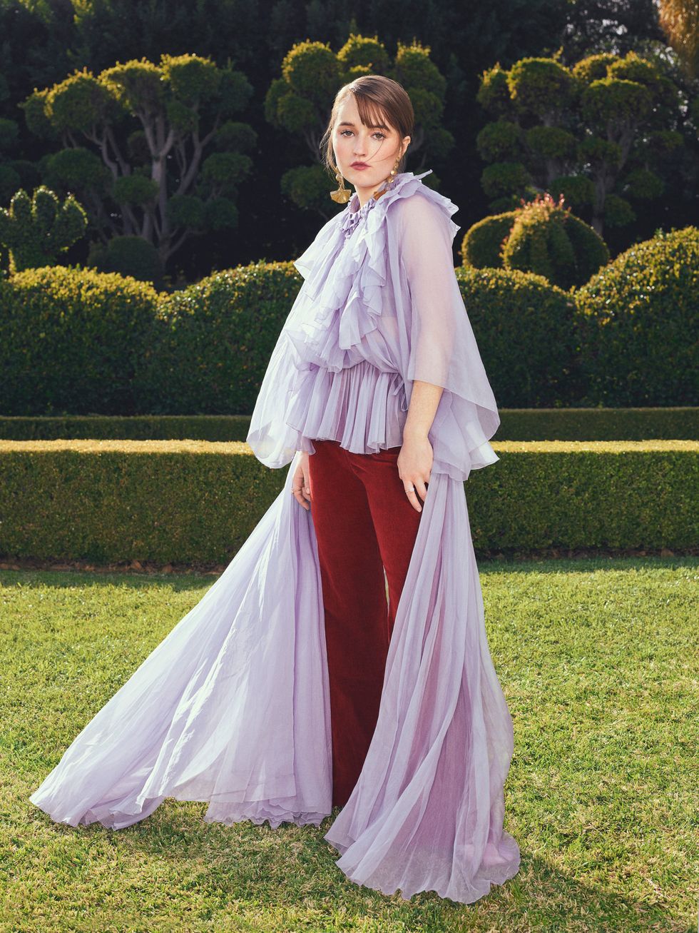 Actress Kaitlyn Dever Stands in a topiary garden wearing burgundy pants and long flowing lilac colored top.