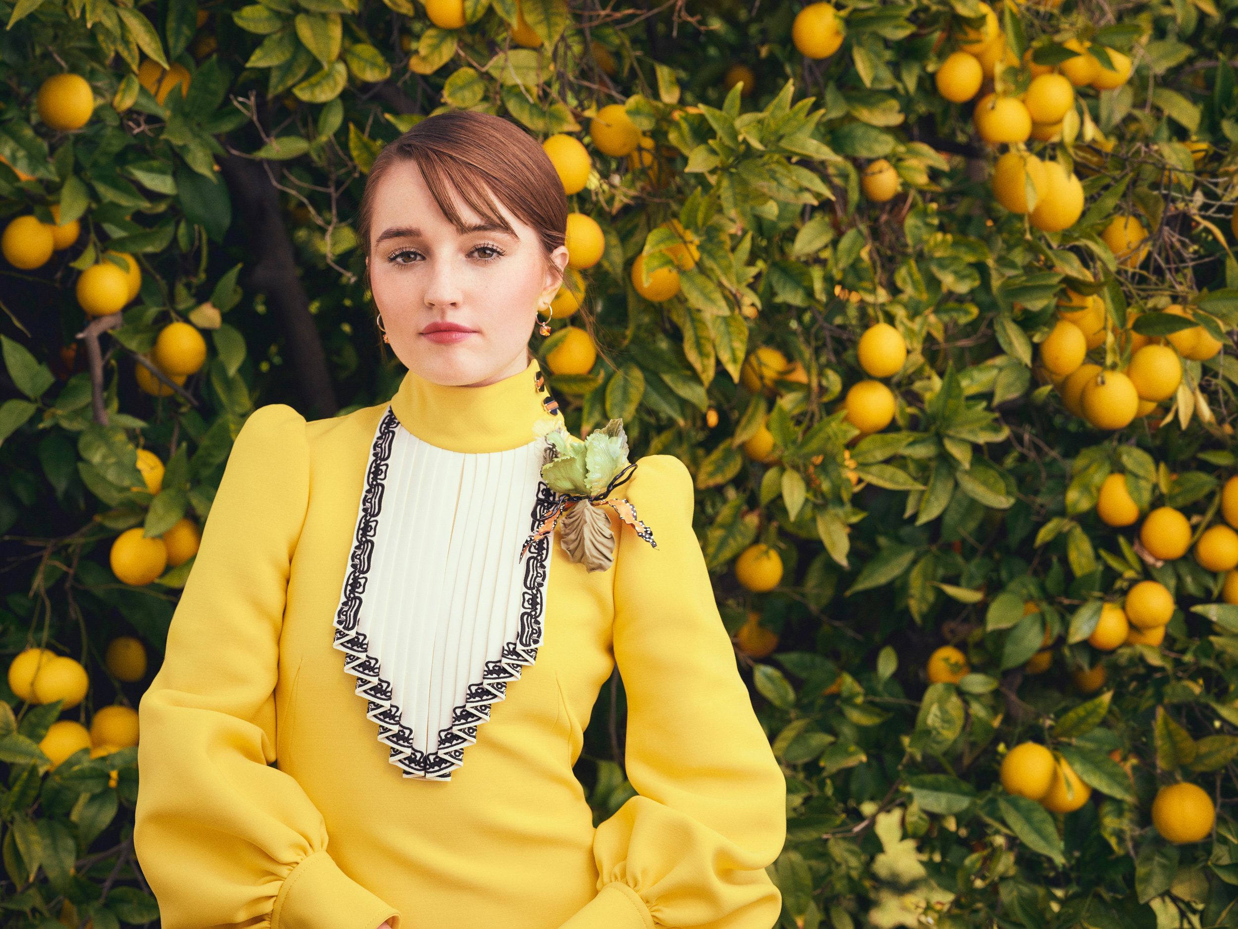 Actress Kaitlyn Dever wears a yellow dress as she stands in front of an orange tree full of fruit.