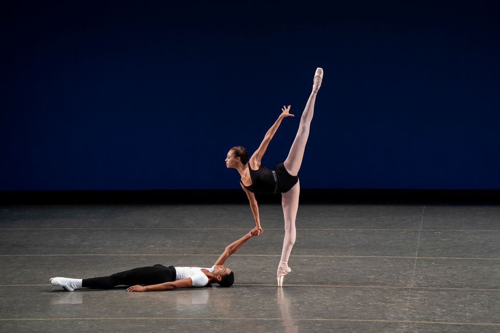 A male dance student in black tights and a white t-shirt lays down on the stage, holding the right hand of a female dance student as she balances in pench\u00e9 on pointe.