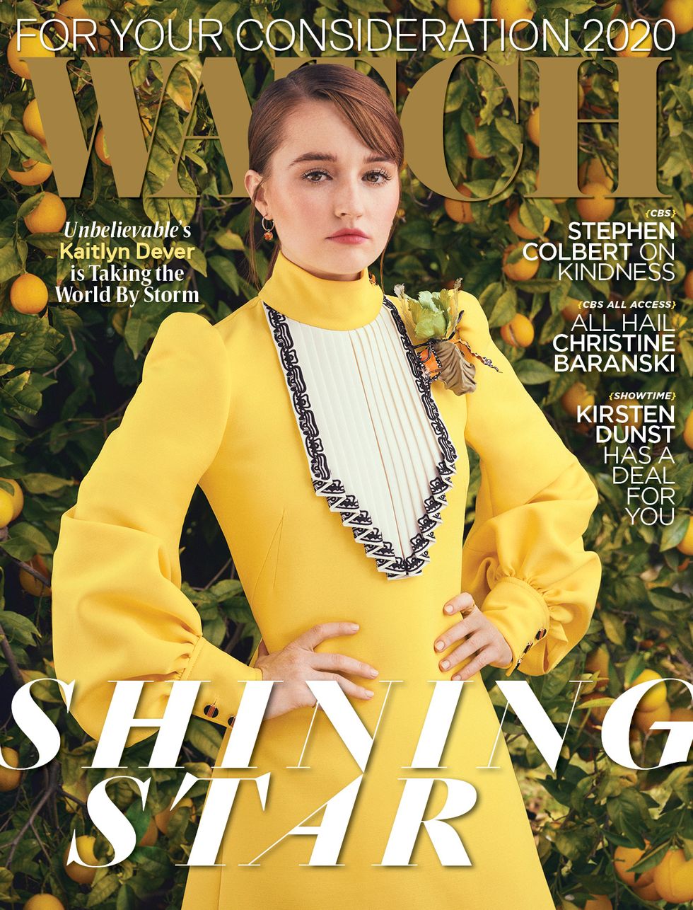 Actress Kaitlyn Dever wears a yellow dress as she stands in front of an orange tree full of fruit.