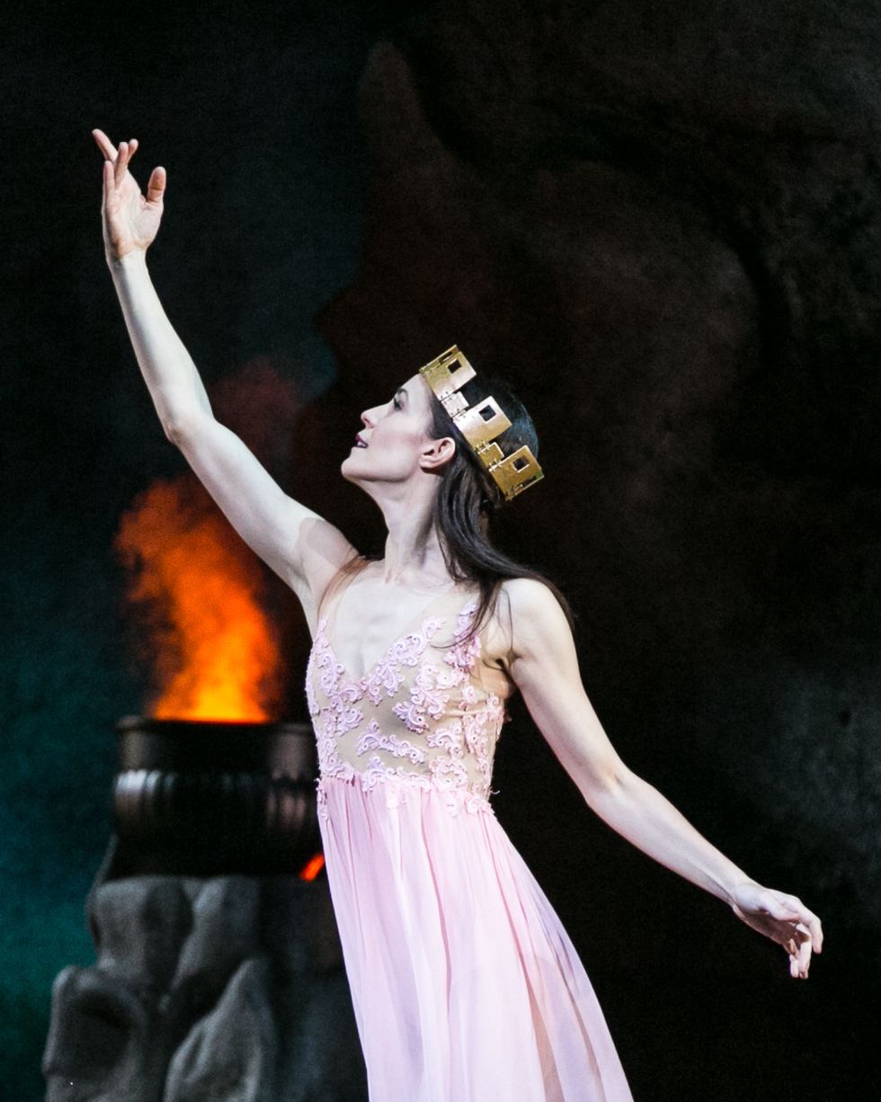 Lara O'Brien, wearing a pink dance dress and a chunky gold crown, makes a beckoning motion with her right arm and looks toward her right.