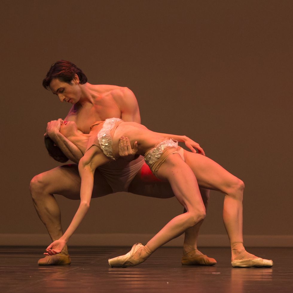 Eris Nezha, wearing white briefs, squats into second position and cradles the head of Petra Conti, who is wearing a sparkly white top and briefs.