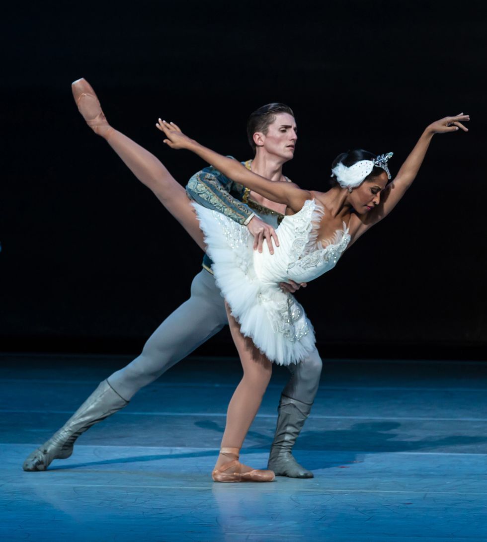 Kayla Rowser, costumed in a white swan tutu, pli\u00e9s in first arabesque allong\u00e9 on her left leg. Nicholas Scheurer, in a blue-green price costume, holds onto her waist behind her.