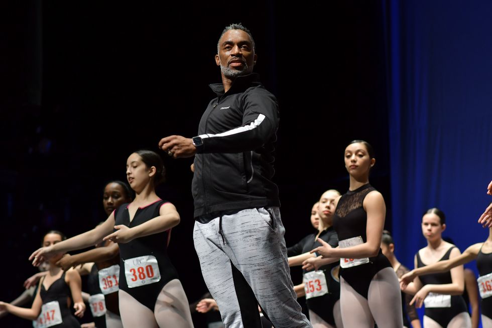 Richardson stands onstage in front of a group of young female dancers in black leotards and pink tights, demonstrating a step.