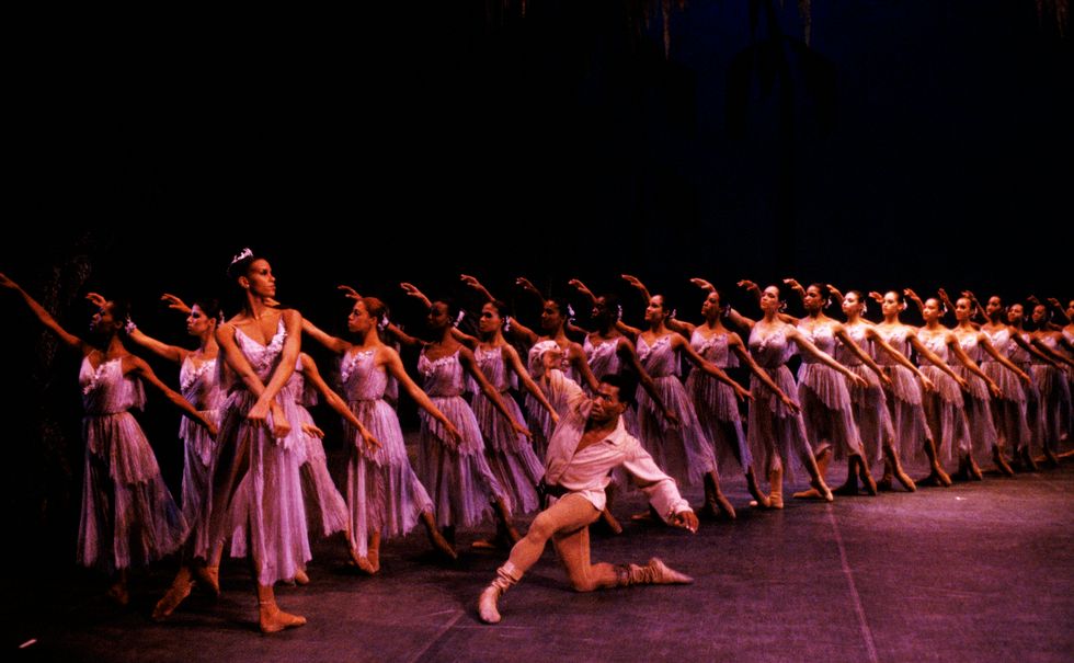 Lorraine Graves, wearing a purple dance dress and crown of flowers, stands on her left leg in tendu derri\u00e9rre with her wrists crossed in front of her. Behind her stand a line of similarly dressed wilis in tendu, while Lowell Smith kneels in front of Graves.