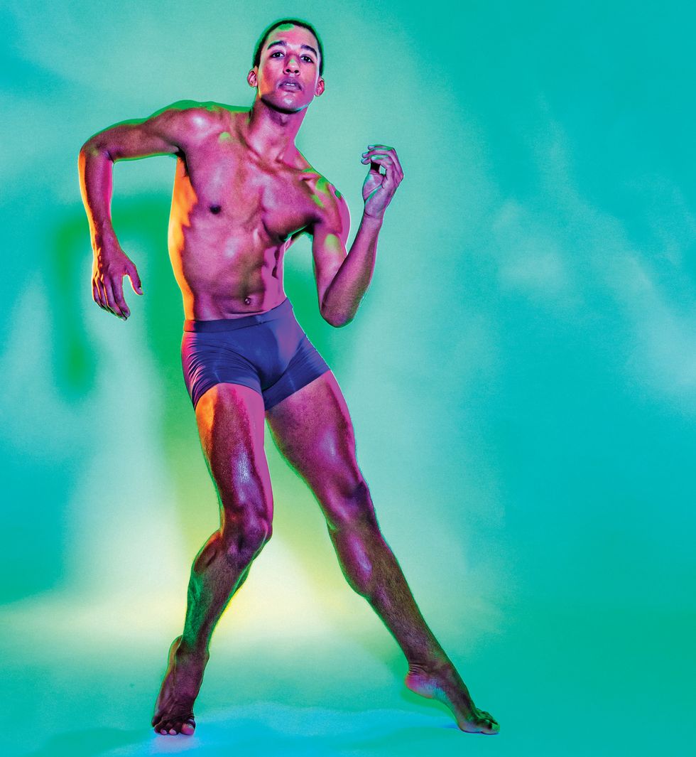 Watters, in purple bike shorts, poses in forced-arch with his arms at an angle, staring directly into the camera, on a teal seamless