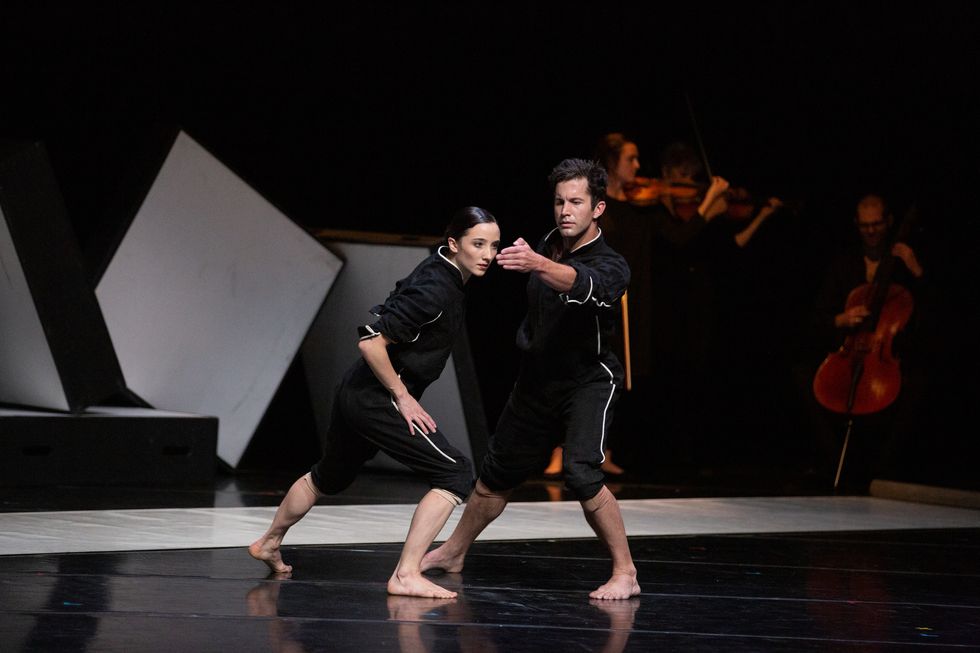 Carly Wheaton and Gabriel Gaffney Smith, wearing knee-length black leggings and zip-up tops, face each other and lunge towards the audience. Wheaton looks into Smith's outstretched hand as a string quartet plays in the background.