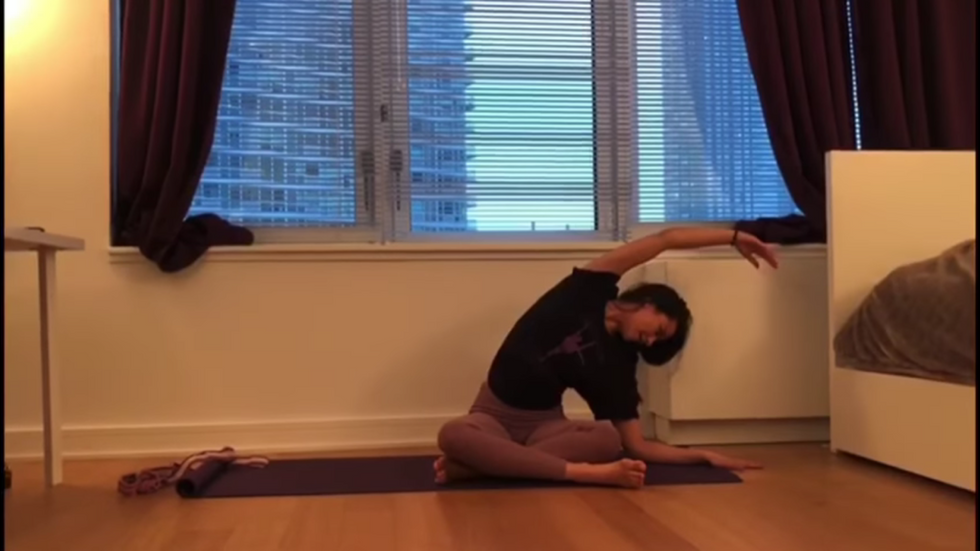 Crystal Serrano wearing warm-up clothes sits cross-legged on a yoga mat, stretching. Her bed is visible to the right.