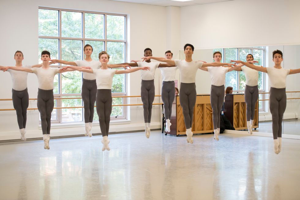 A group of 10 boy ballet students wearing white T-shirts and gray tights jump in soubresaut, with their arms in second position all facing front.