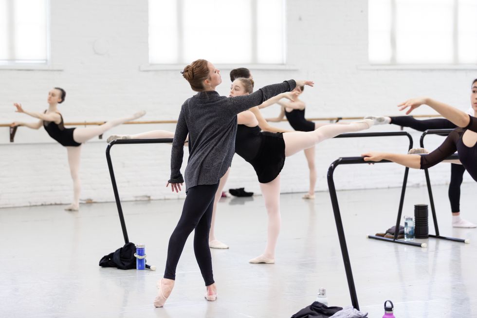 A ballet teacher in black leggings, a gray sweatshirt and ballet shoes stands in tendu derriere with her right arm stretched in front of her while a class of female ballet students do a similar position in second arabesque.