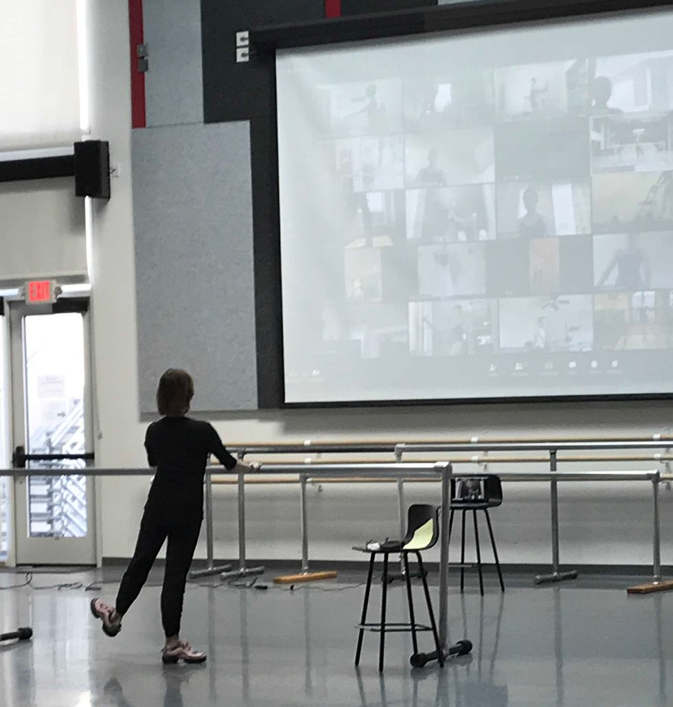 Standing with her back to the camera, a teacher in all black practices tendu side with a flexed foot, facing a large screen showing her students following along on Zoom.