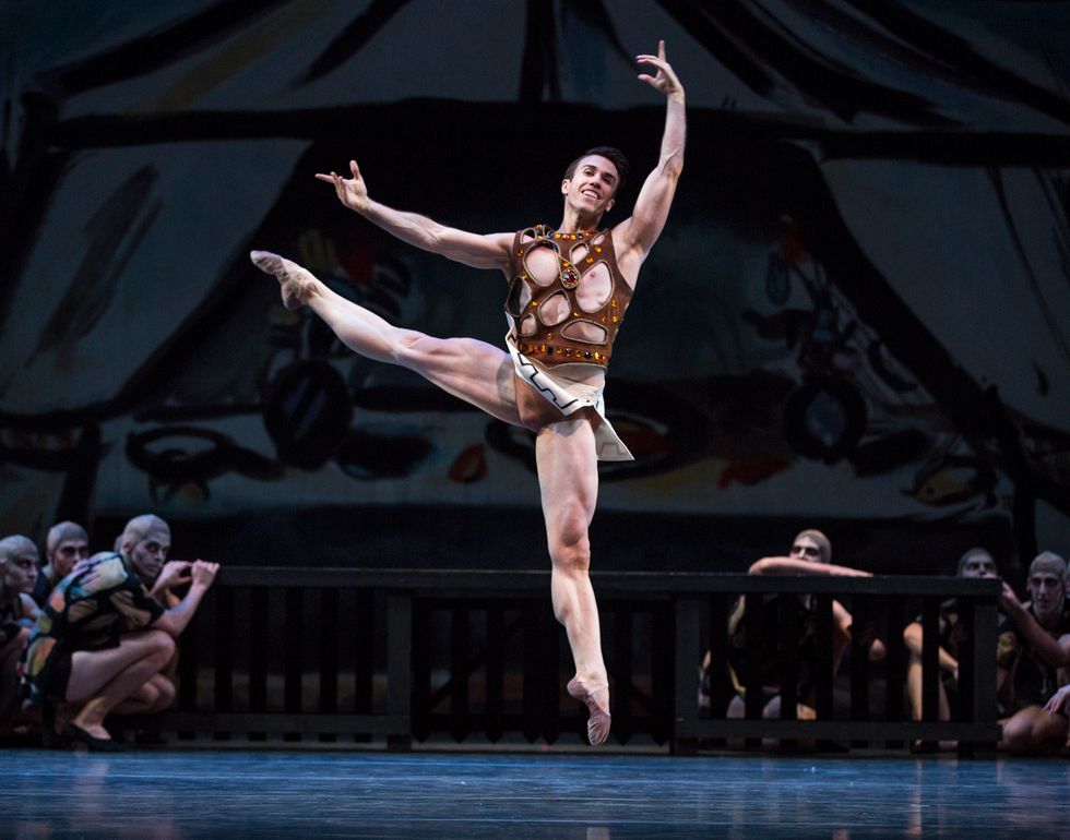 Benjamin Griffiths, wearing a brown shirt and short off-white skirt, jumps off of his left leg with his right leg held high the side and his left arm held over his head.