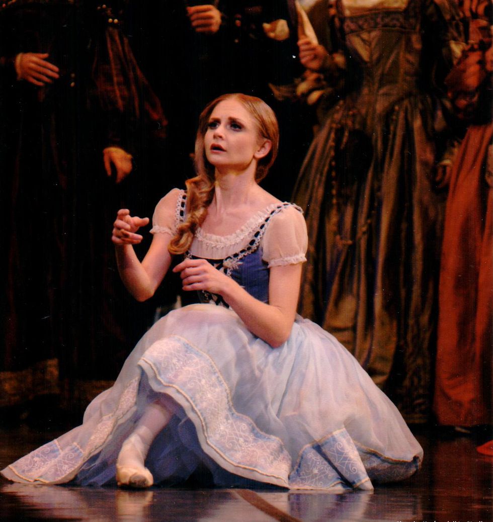 Chandra Kuykendall, dressed in a blue peasant dress and with her blonde hair down, sits on the stage floor and looks far off into the distance.