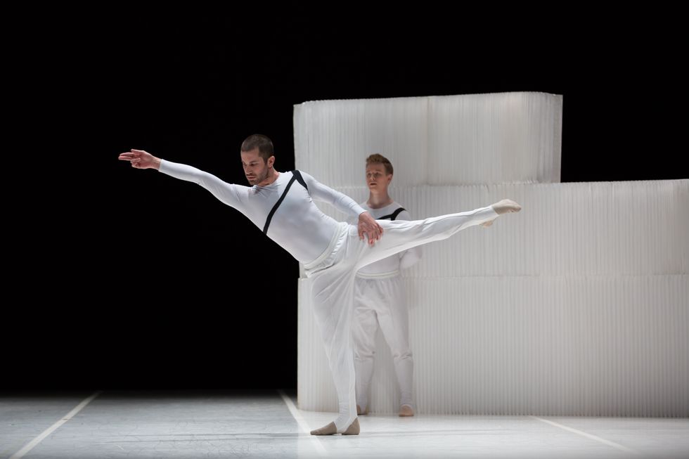 Gabriel Gaffney Smith, wearing a white shirt and stirriped pants, stands on his right leg in a twisted open first arabesque. Behind him stands a similarly dressed male dancer in front of a large, white rectangular structure.