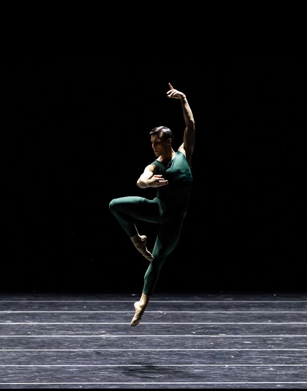 Wearing a green unitard, Cameron Thomas performs a saut\u00e9 jump with his right leg in retir\u00e9 and his left arm held above his head.