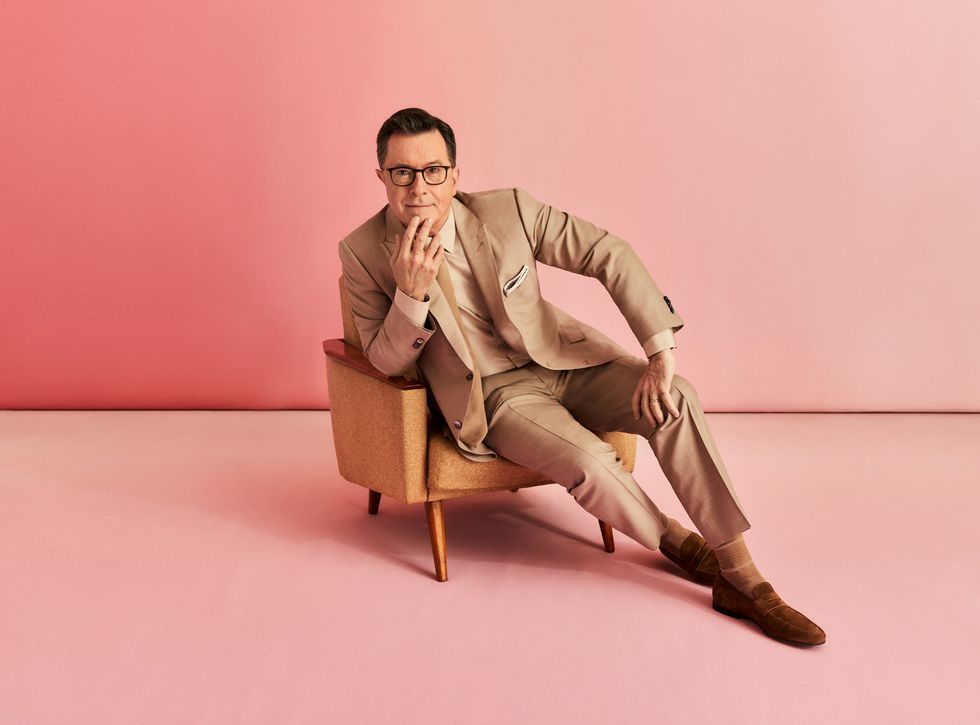 Stephen Colbert sitting in a chair against a pink backdrop.