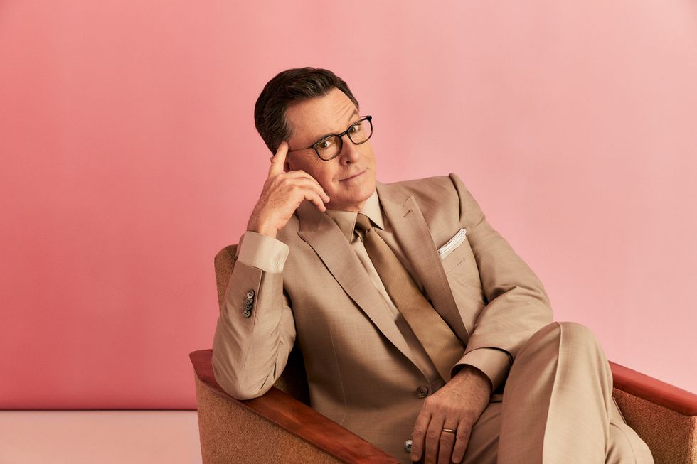 Stephen Colbert sitting in chair in a tan suit.