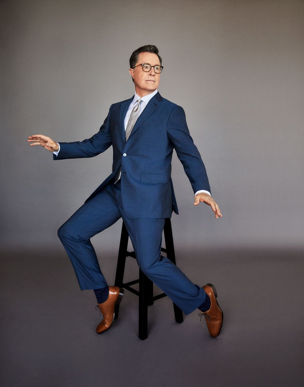 Stephen Colbert in a navy blue suit sitting on a stool.