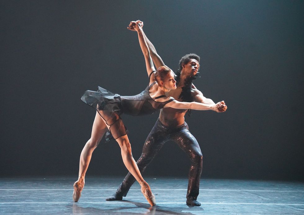 Jillian Davis, in a black leotard and short, contemporary tutu, stands in fourth position on pointe and reaches over at the waist while holding Kayr Muhammad's hands in a stretched port de bras.