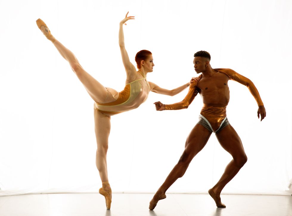 Jillian Davis, wearing a gold leotard, stands in arabesque on pointe. She balances her left hand on Brandon Gray's shoulder and holds her right arm above her head while he poses in tendu second with his left foot in demi-pointe.