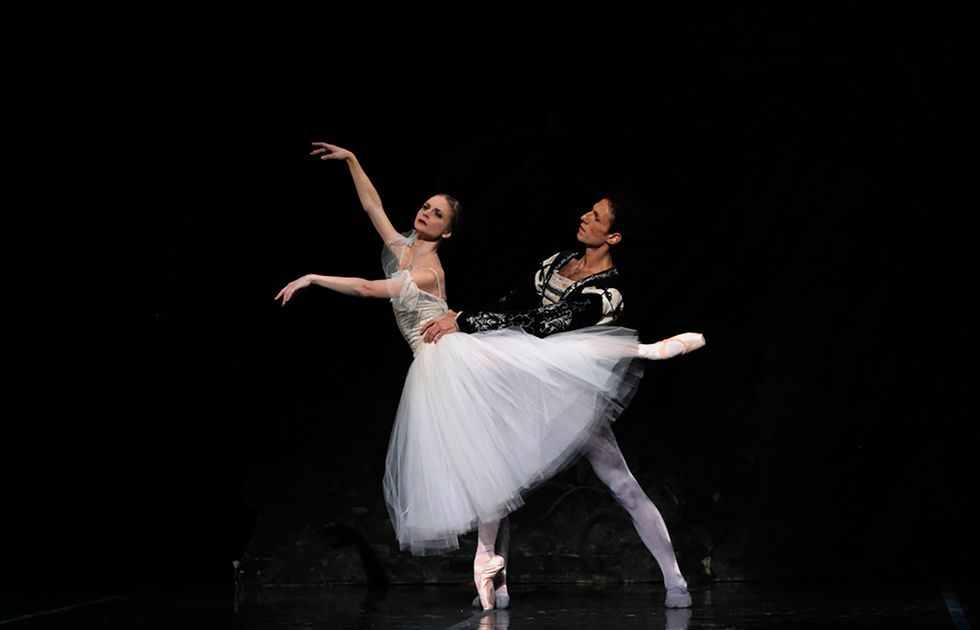 Chandra Kuykendall, wearing a white Romantic tutu, poses in third arabesque on her right leg while Domenico Luciano, in a black and white tunic and gray tights, stands behind her on her left, holding her waist.