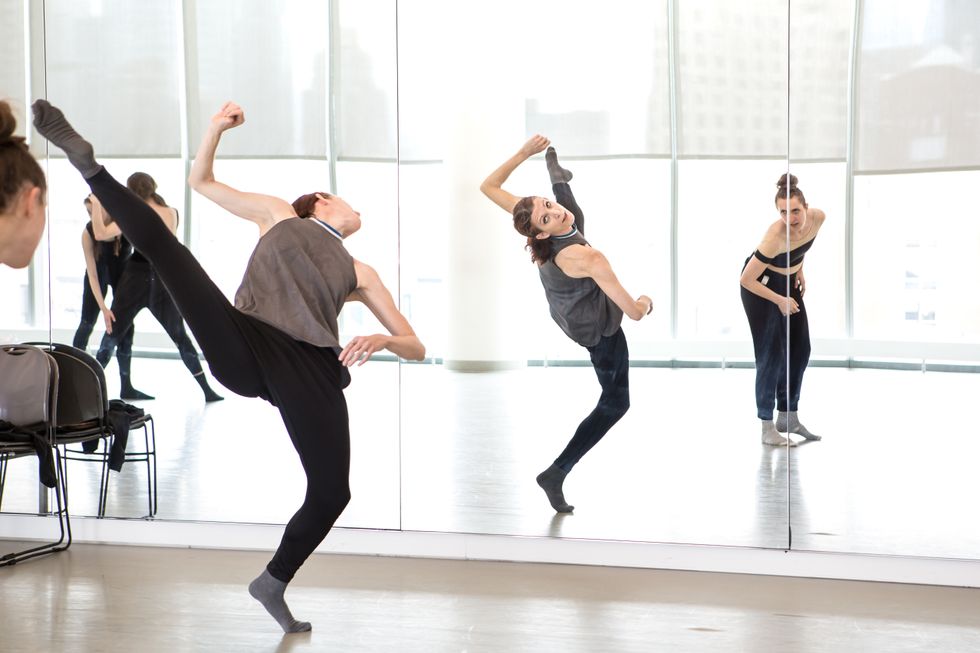 Wearing black leggings and sleeveless gray top, Gabrielle Lamb kicks her right leg high and twists her upper body back, looking at her reflection in the mirror. Three dancers in black dancewear look on.