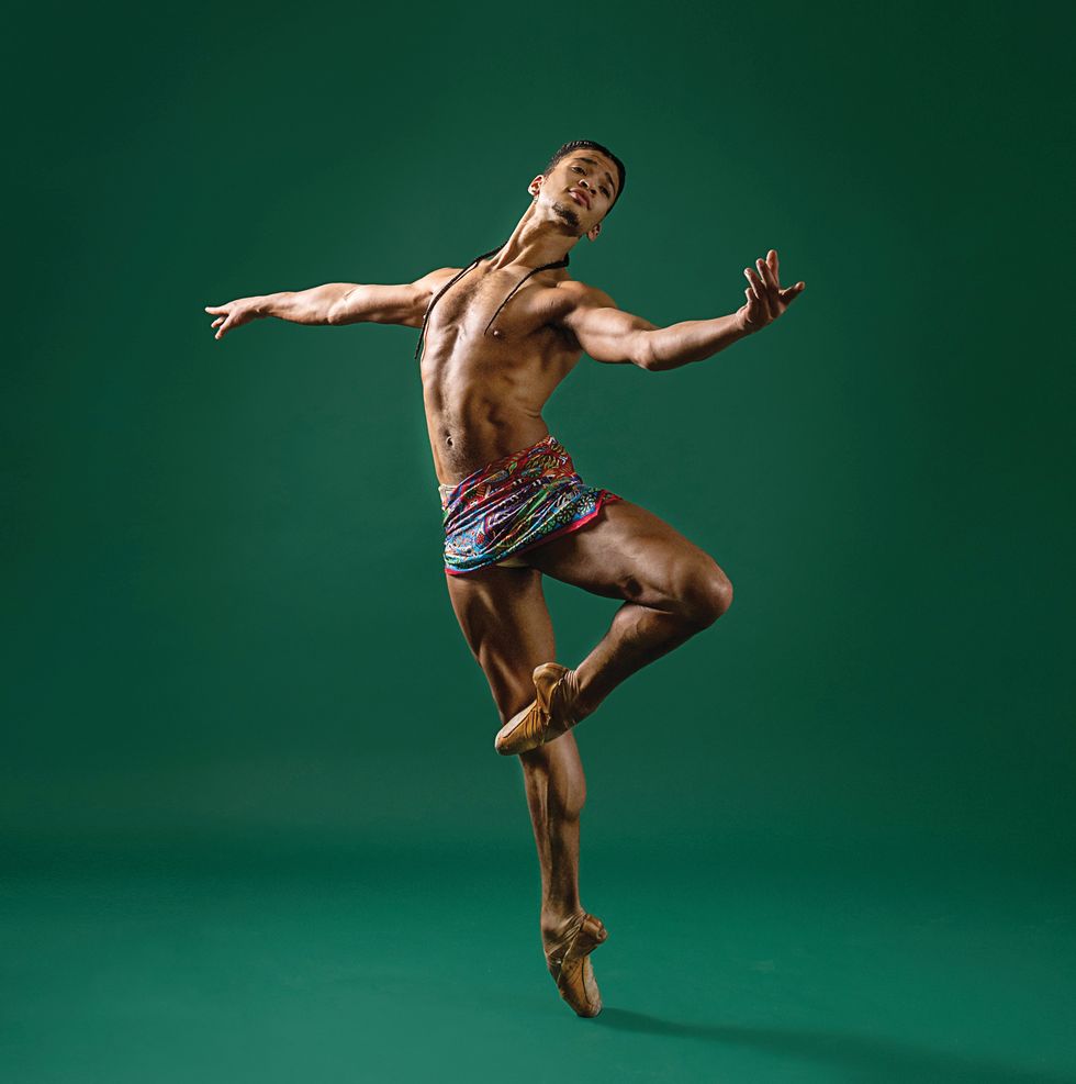 Hayes in a short colored wrap skirt and pointe shoes in a pass\u00e9 with his arm extended to the side