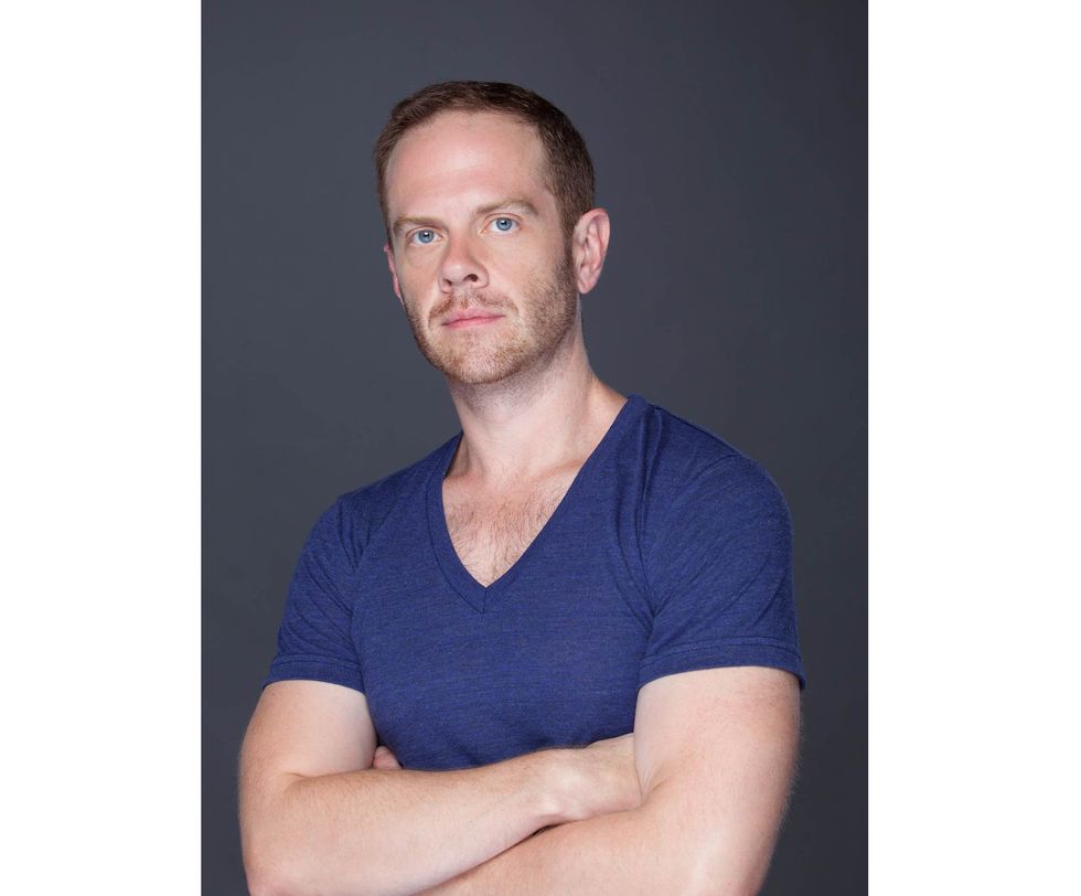 Matthew Neenan, wearing a blue V-neck T-shirt, is shown from the waist up crossing his arms and looking towards the camera.