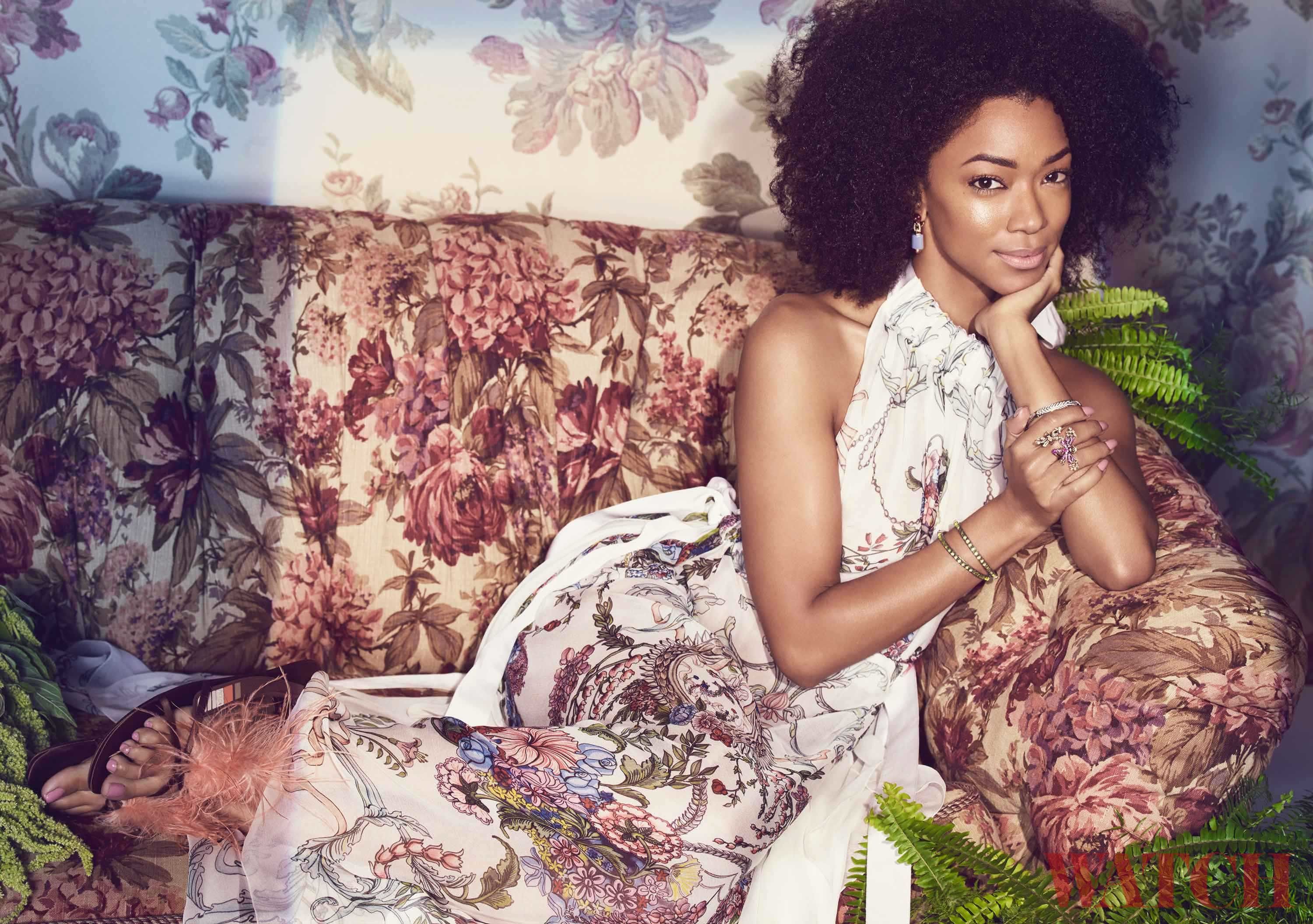 Sonequa Martin-Green reclines a floral print couch in a white patterned dress.