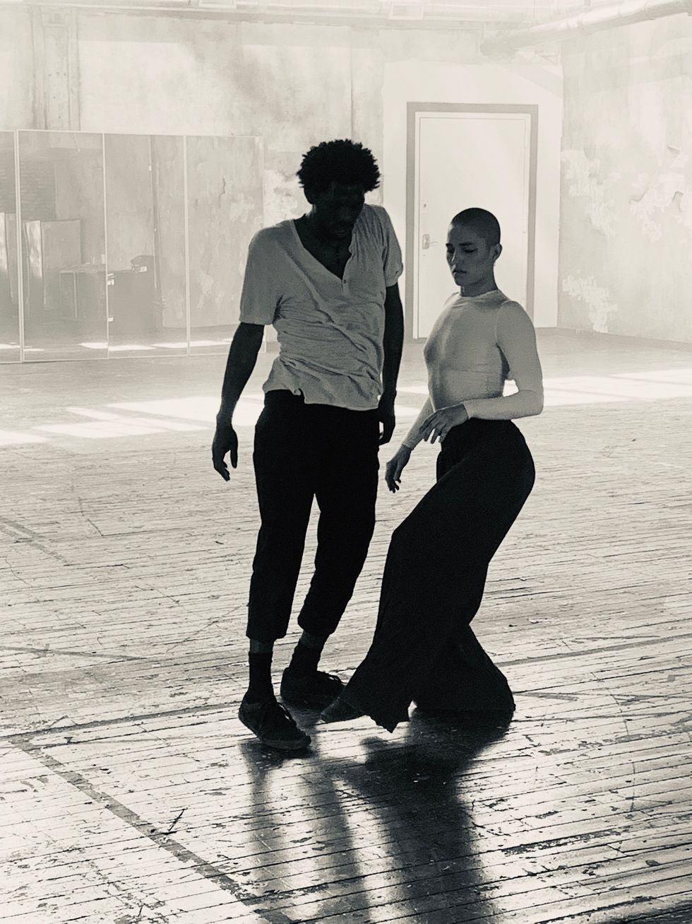 Black and white photo of two dancers moving close together but not touching in an empty warehouse-like room.