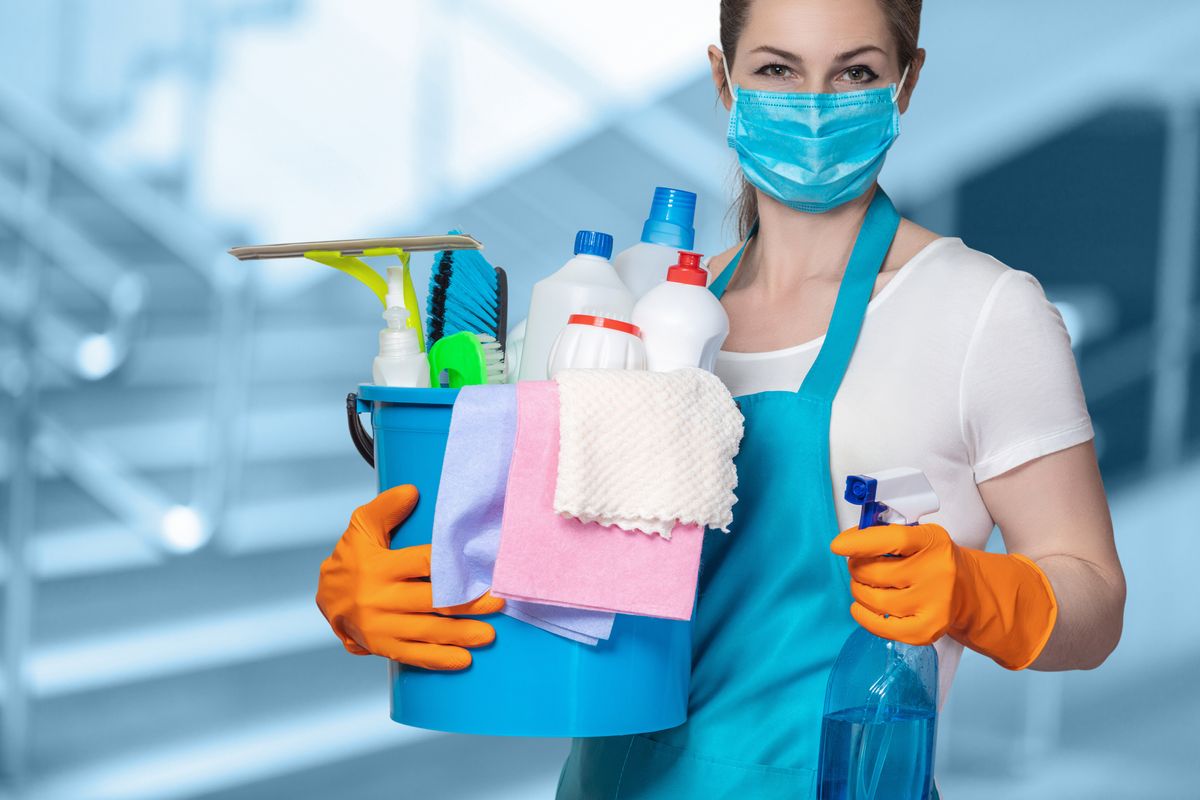woman in a blue face mask holds a bucket of assorted generic cleaning products