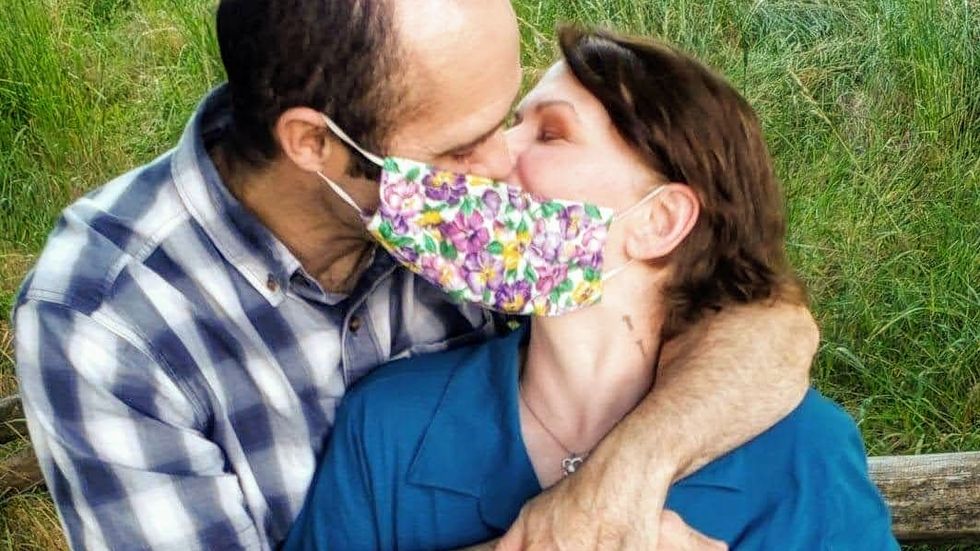 A man and a woman kissing with a flower-printed mask hooked on one of his ears and one of her ears, so that the mask covers their mouths