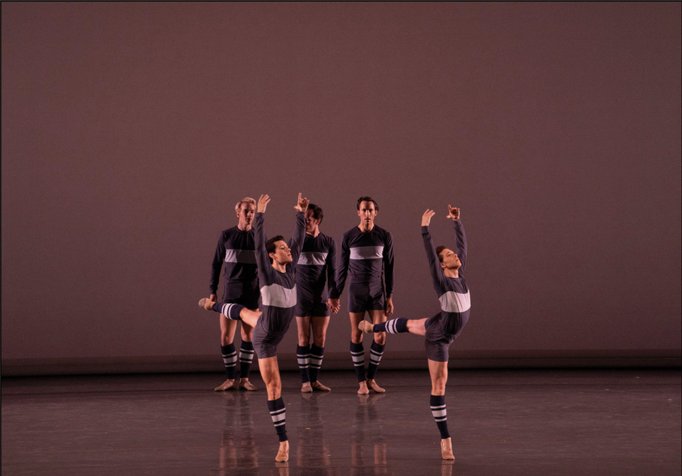 Two male dancers in blue shorts, a blue long-sleeved shirt with a large gray stripe, and blue and gray striped legwarmers, do piqu\u00e9 arabesque with their arms in high fifth. Three men in similar costumes stand casually behind them onstage.