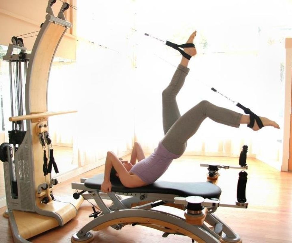 Wearing workout clothes, Charlene Aldred lays on a Gyrotonic reformer with her legs scissoring in the air and her feet in foot straps.