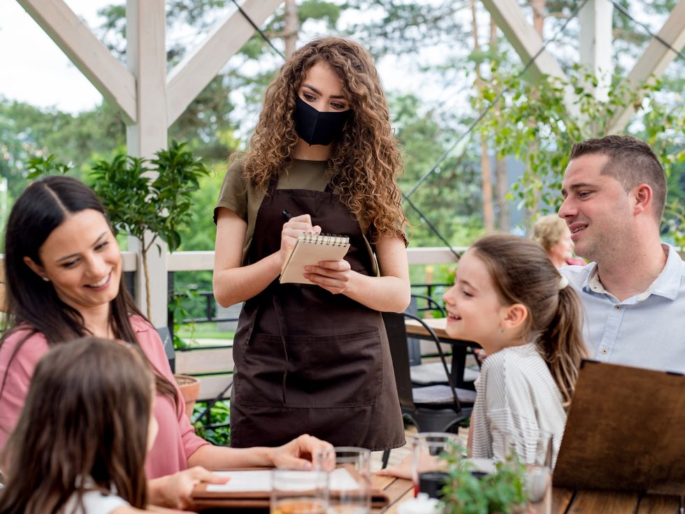 Waitress with face mask serving family with children outdoors in summer on terrace restaurant