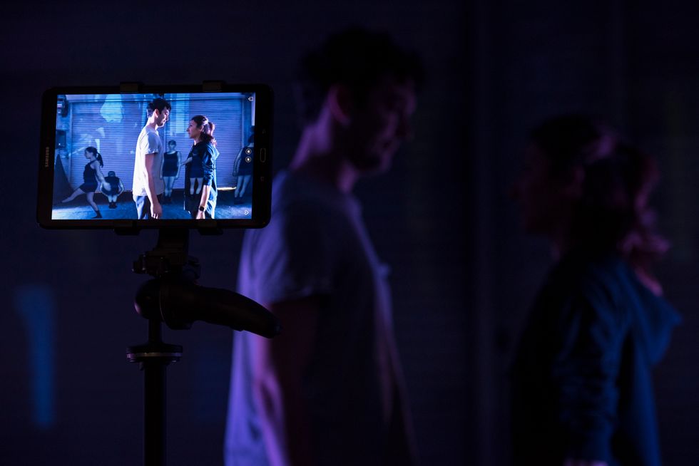 In a darkened space, an iPad on a tripod shows a male and female dancer, who are in the background behind the device, in street clothes facing each other.