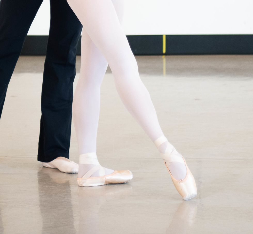 A young female dancer is shown in tendu crois\u00e9 devant from the knee down, wearing pink tights and pointe shoes.