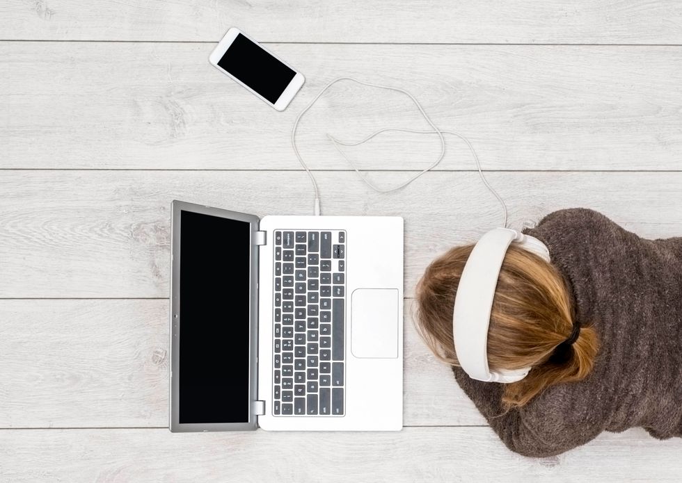 A pony-tailed girl wearing a brown sweater and white headphones sits on the floor and looks at her laptop and smartphone.
