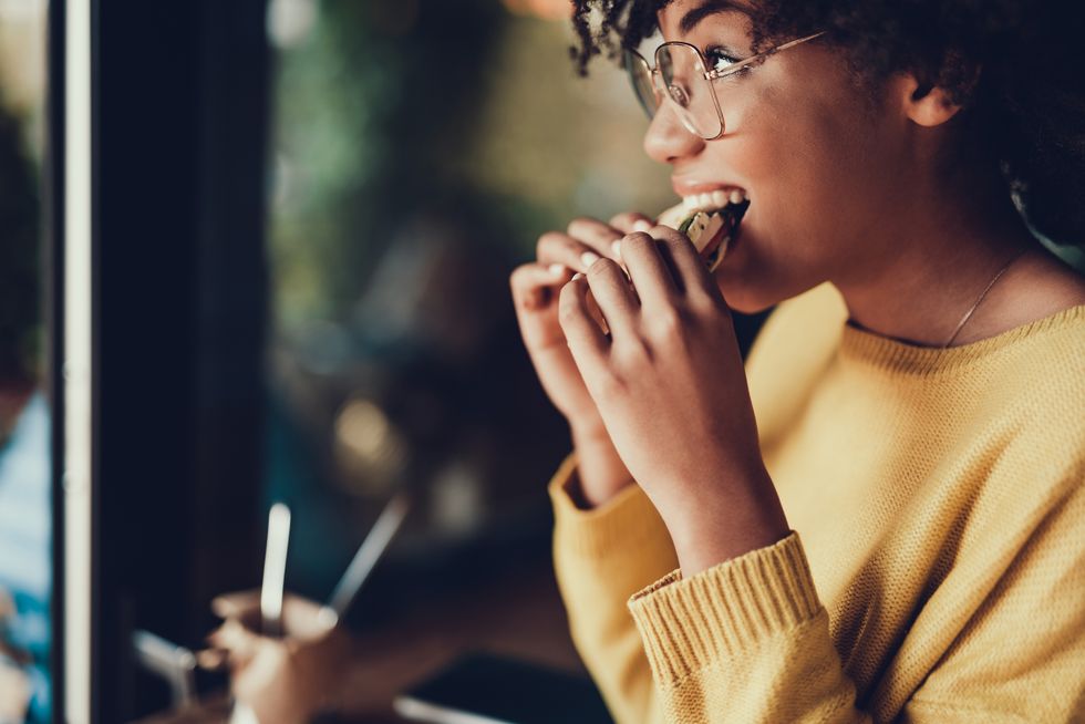 A black woman in a mustard sweater and glasses bites into a sandwich.
