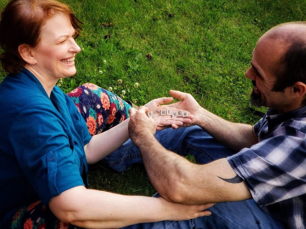 The author and her fiance sitting on the grass with the word "forever" spelled in Scrabble letters on her hand with her ring for the "o"