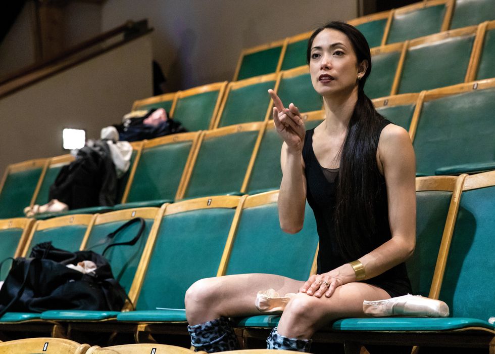 Abrera sits in a green theater seat, pointing at the stage while sewing pointe shoes in her lap.