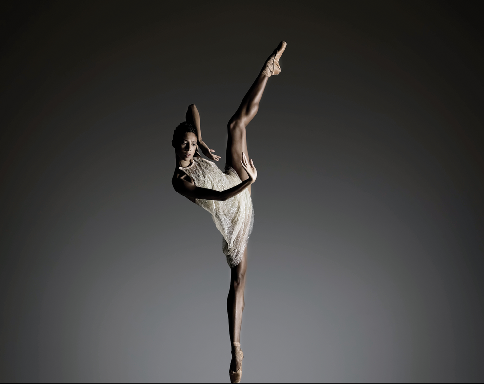 Cissoko, in a pale sparkly dress and pointe shoes, extends one leg high in the air in a modern position against a gray background.