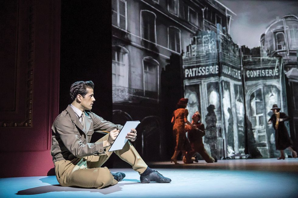 Fairchild, dressed in slacks and a brown jacket and tie, sits on the stage with a sketchpad while dancers move around behind him in a Parisian street set